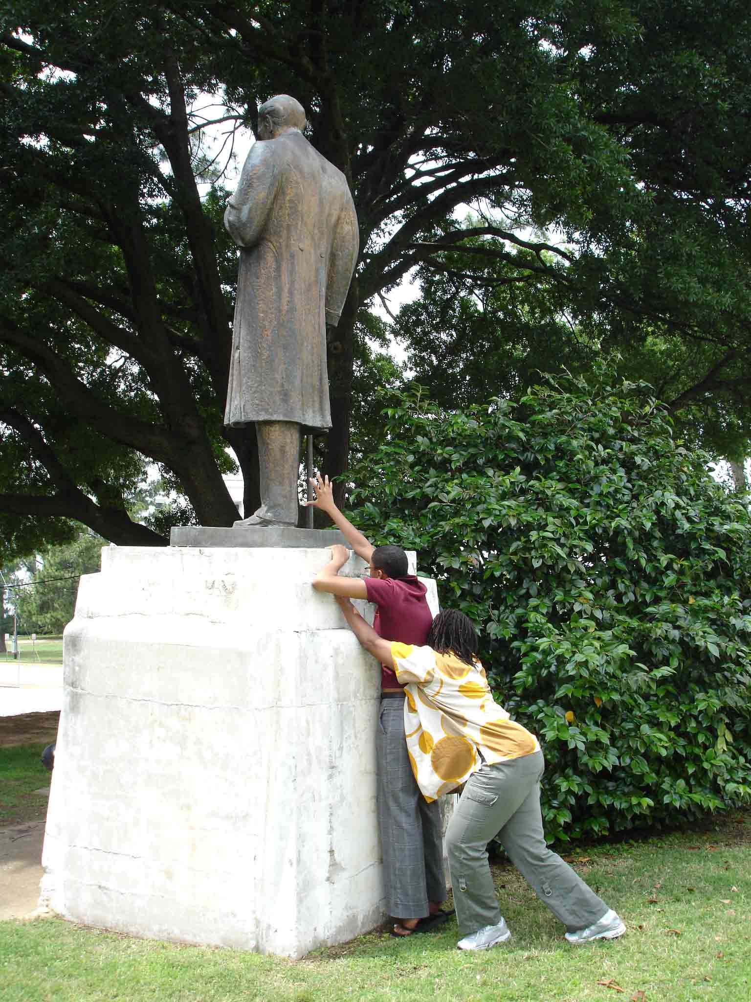 Tabitha and Anita reaching up to the statue of Sims, a huge monument with its back turned towards them